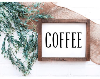 May include: A wooden framed sign with the word "COFFEE" in bold black letters on a white background. The sign is set against a white brick wall, with green leafy vines and a burlap cloth in the background.
