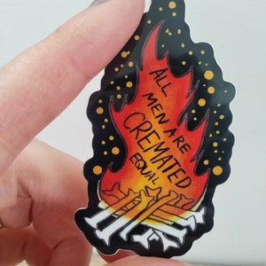 May include: A black and red sticker with a flame design. The flame has a yellow and orange gradient. The text "ALL MEN ARE CREMATED EQUAL" is written in white on the flame. There are small yellow dots around the flame.