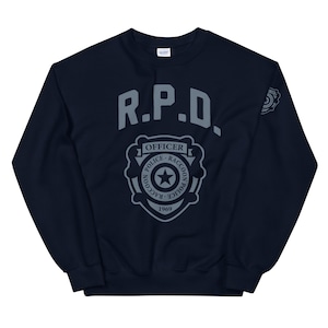 Könnte beinhalten: Marineblaues Sweatshirt mit grauem R.P.D.-Officer-Abzeichen. Das Abzeichen enthält den Text "Raccoon Police 1969" und einen Stern. Ein kleineres Abzeichen befindet sich auf dem rechten Ärmel.