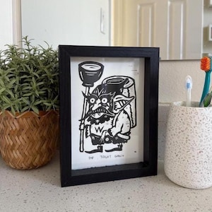Pode incluir: Impressão artística emoldurada em preto com uma ilustração humorística de um goblin segurando um desentupidor e papel higiênico. O texto "The Toilet Goblin" está impresso abaixo. A obra de arte é exibida em uma bancada de banheiro.