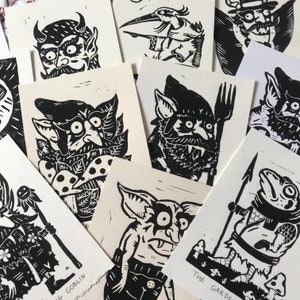 May include: A collection of black and white linocut prints featuring various goblin and creature illustrations. The prints are on white cardstock and have a hand-carved, rustic aesthetic. Some of the prints include text, such as "The Garden" and "Goblin".
