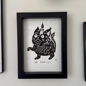 May include: Black and white framed art print featuring a stylized cat with a candle on its head. The cat has a stern expression and is labeled "The Night Cat" in a bold font. The frame is black.