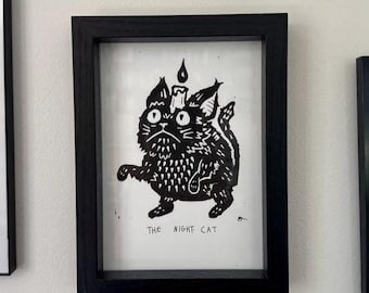 Può includere: Stampa incorniciata in bianco e nero raffigurante un gatto stilizzato con una candela sulla testa. Il gatto ha un'espressione seria e il testo "The Night Cat" è in basso. La cornice è nera.