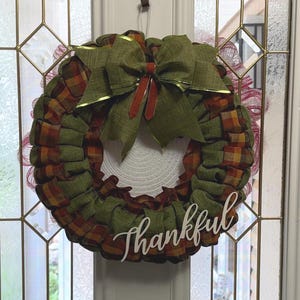 May include: A circular wreath featuring alternating loops of olive green fabric and orange, brown, and yellow plaid fabric. A large olive green bow sits at the top, and the white cursive text "Thankful" is attached to the lower right side.