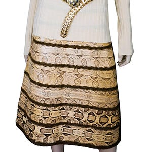 May include: A beige skirt with a snakeskin pattern and dark brown horizontal stripes. A gold-colored belt with a decorative buckle is worn at the waist. The model is wearing a cream-colored long-sleeved top.