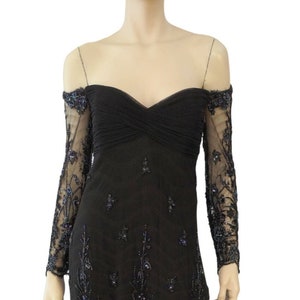 May include: A black off-the-shoulder gown with long sleeves. The sleeves are made of sheer fabric with beaded floral embroidery. The bodice is fitted and the skirt is flowing.