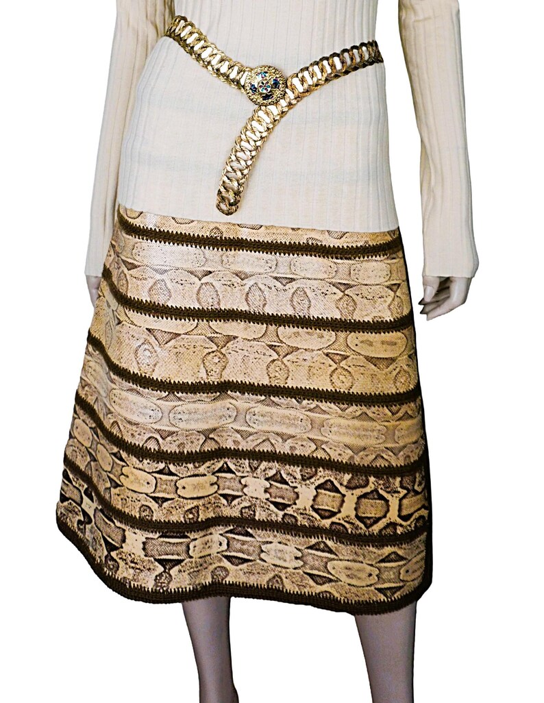 May include: A beige skirt with a snakeskin pattern and dark brown horizontal stripes. A gold-colored belt with a decorative buckle is worn at the waist. The model is wearing a cream-colored long-sleeved top.