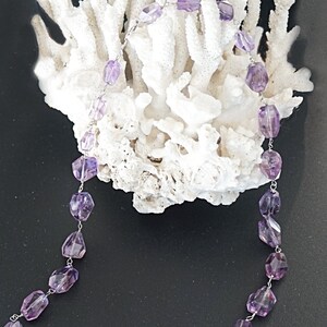 May include: A purple amethyst gemstone necklace with faceted beads and silver chain links. The necklace is draped over a white coral branch.