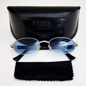 Puede incluir: Gafas de sol con montura plateada y lentes azules, un estuche negro con la inscripción "FENDI" y un paño de limpieza negro. Las gafas de sol tienen una forma ovalada clásica y patillas negras. El estuche es brillante y rectangular.