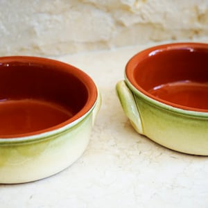 May include: Two terracotta bowls with green and yellow glaze. The bowls have rounded edges and two small handles on each side.