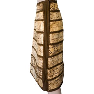 May include: A knee-length skirt with a snakeskin pattern in beige and brown tones. The skirt is constructed from rectangular panels, with dark brown knitted trim and stitching. The skirt is displayed on a mannequin.
