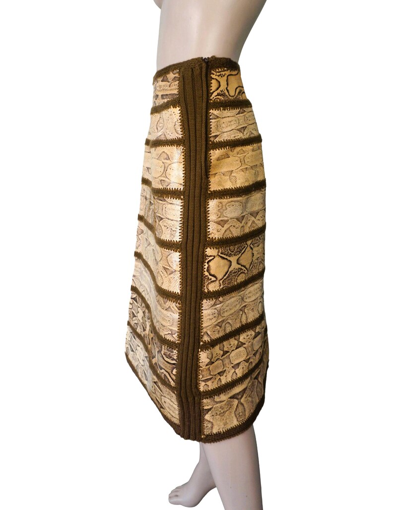 May include: A knee-length skirt with a snakeskin pattern in beige and brown tones. The skirt is constructed from rectangular panels, with dark brown knitted trim and stitching. The skirt is displayed on a mannequin.