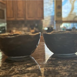 May include: Two small, dark brown ceramic bowls with irregular, wavy rims and textured, dimpled outer surfaces. The bowls have a glossy finish and a white base visible at the bottom, showcasing a handcrafted appearance with organic, uneven shapes.