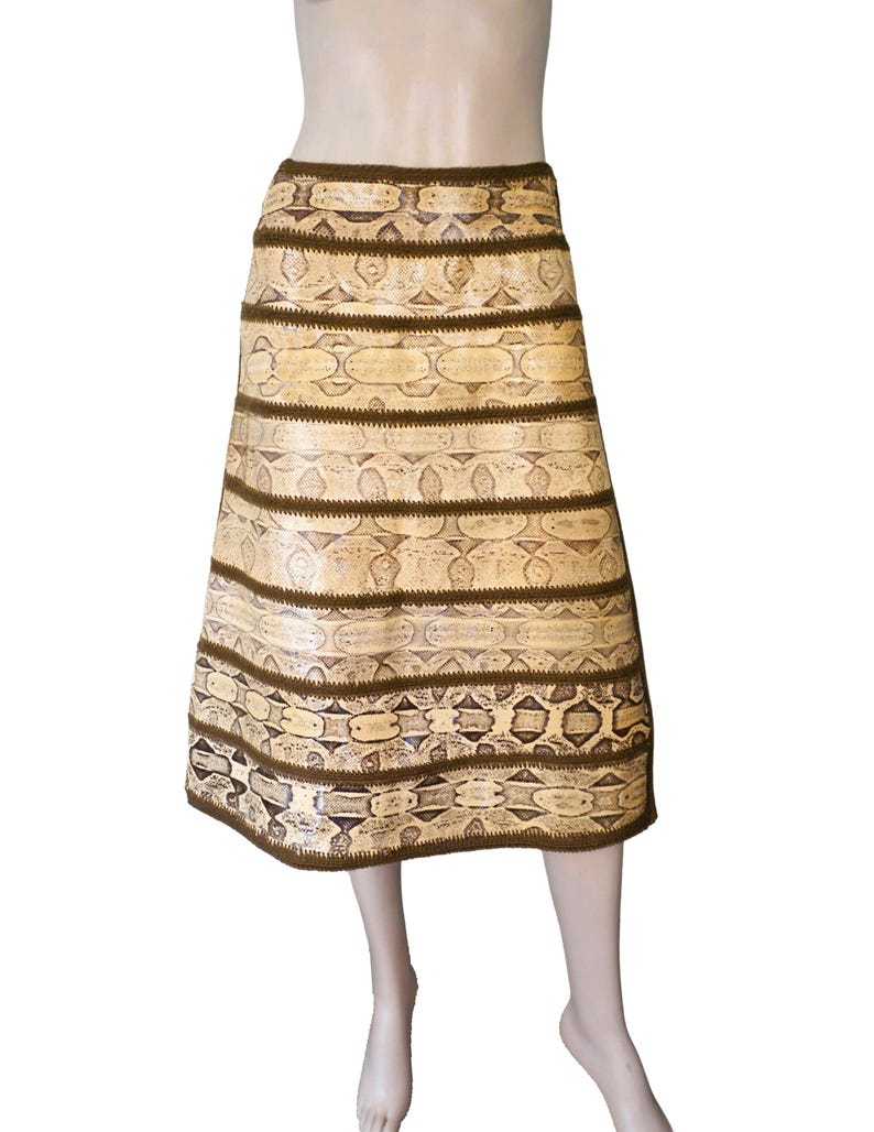 May include: A knee-length skirt with a snakeskin pattern in shades of beige and brown. The skirt features horizontal bands of the snakeskin pattern separated by dark brown trim. The skirt is displayed on a mannequin.