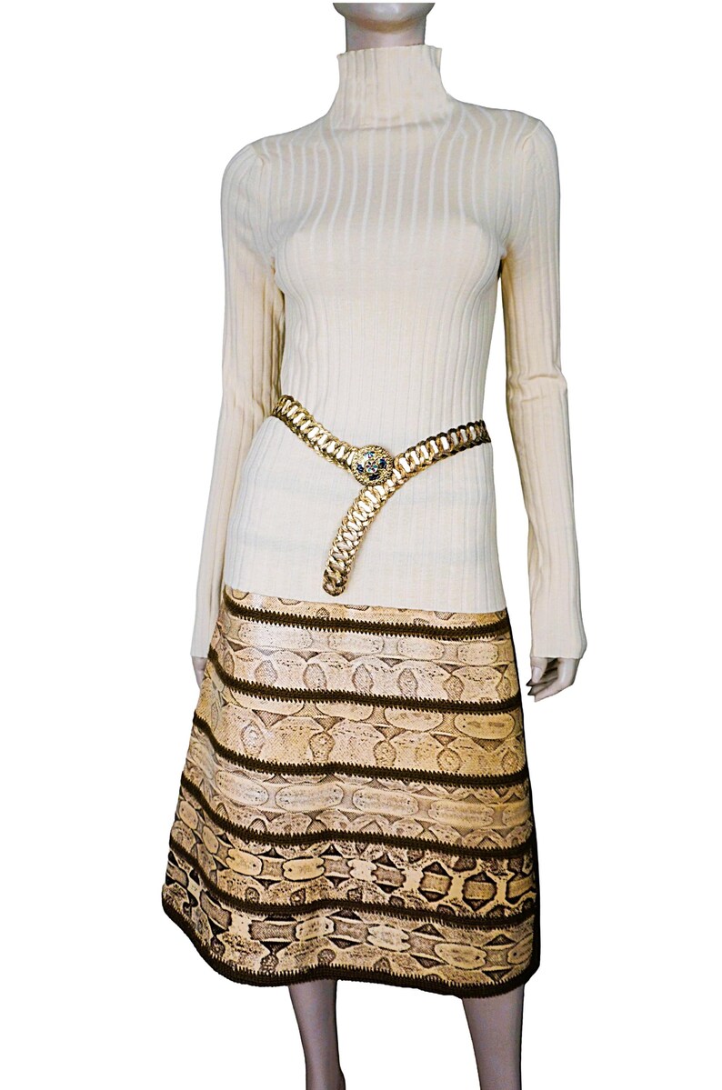 May include: A cream-colored turtleneck sweater paired with a gold chain belt and a snakeskin-patterned skirt. The belt features a decorative center piece. The skirt has horizontal bands of brown and tan patterns.