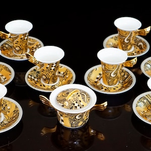 May include: A set of eight gold and white porcelain coffee cups and saucers with a decorative pattern. The cups have a unique wing-shaped handle. There is also a matching sugar bowl with a lid.