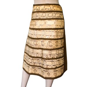 May include: A knee-length skirt with a snakeskin pattern in shades of beige and brown. The skirt features horizontal bands of the snakeskin pattern separated by dark brown trim. The skirt is displayed on a mannequin.