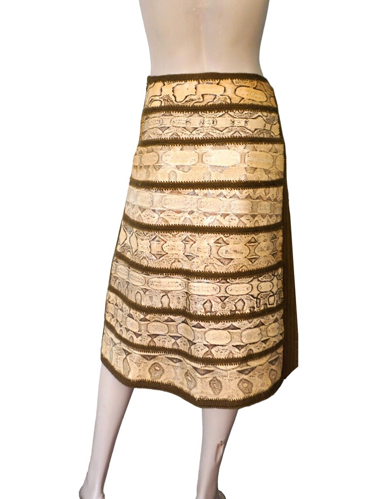 May include: A knee-length skirt with a snakeskin pattern in shades of beige and brown. The skirt features horizontal bands of the snakeskin pattern separated by dark brown trim. The skirt is displayed on a mannequin.