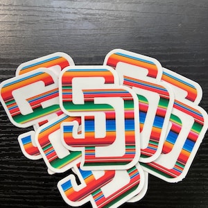 May include: A stack of white stickers with a white letter "S" inside a colorful striped pattern. The stripes are red, orange, yellow, green, blue, and purple.