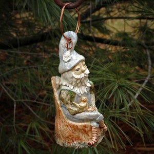 May include: A ceramic gnome figurine with a blue hat and white beard, sitting on a tree stump and holding a bird in its hand. The gnome is wearing a green shirt and blue trousers.