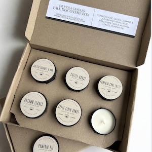 May include: A brown cardboard box containing six small round soy candles. The lid features the text Val Viola Candles Fall Discovery Box and lists scents including Coffee House, Salted Caramel & Oud, Pumpkin Chai Latte, Apple Cider Donut, Autumn Leaves, and Pumpkin Pie. Each candle has a circular label.