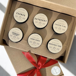 May include: A gift box containing six soy wax candles in small, round tins. Each tin has a black lid and a label with a scent name like "Eggnog" and "Bayberry." The box is tied with a red ribbon.