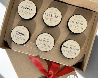 May include: A presentation box of six soy wax candles in small, round tins. Each tin has a black lid and a label with a scent name such as "Eggnog" and "Bayberry." The box is tied with a red ribbon.