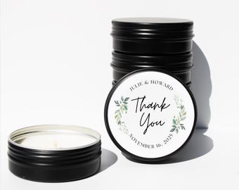 May include: Black metal candle tins, one open, with a white candle and wick. The lids have a white label with "Thank You", names "Julie & Howard" and date "November 16, 2023", surrounded by green floral design. Ideal for wedding favours.
