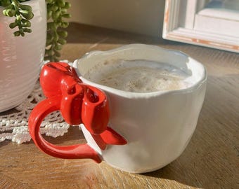 May include: A white ceramic mug filled with a frothy beverage. The mug features a bright red bow-shaped handle. The mug sits on a wooden surface, with a glimpse of a white planter and framed artwork in the background.