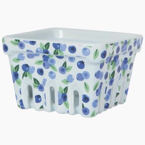 May include: A square white ceramic berry basket featuring a repeating pattern of blue blueberries and green leaves. The container has a wide rim and vertical cutouts along the bottom edges, mimicking the design of a traditional produce carton.