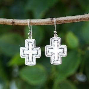 May include: A pair of silver cross earrings with a detailed design. The earrings are hanging from a branch.