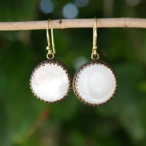 May include: A pair of drop earrings featuring round, iridescent white mother-of-pearl cabochons set in dark, textured bronze-toned metal bezels. The earrings hang from simple gold-toned wire hooks, showcasing a circular design with a polished finish.