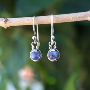 May include: A pair of silver drop earrings featuring round, mottled blue and white stone cabochons. Each earring has a curved silver hook wire and a decorative metal setting that holds the circular stone in place, creating a dangling design.