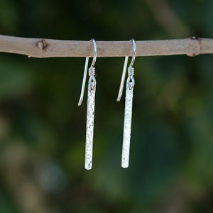 May include: A pair of silver bar earrings with a textured surface, hanging from a thin branch. The earrings have a simple, minimalist design with a hook closure. The background is a soft, blurred green.