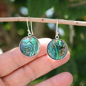 May include: A pair of round earrings with iridescent abalone shells. The earrings have a silver-colored border and a hook closure. The abalone shells display a mix of green, blue, and purple hues, creating a natural, textured pattern.