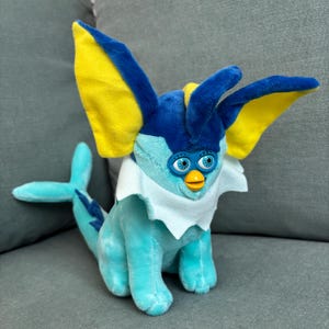 May include: A plush toy resembling a blue and yellow creature with large ears and a tail. The toy has a white collar and blue eyes. The toy is sitting on a grey surface.