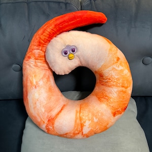 Könnte beinhalten: Ein skurriles Kissen in Form einer riesigen Garnele. Das Kissen hat ein realistisches orange-weißes Garnelenmuster mit einem Furby-ähnlichen Gesicht in der Mitte. Ein einzigartiger und verspielter Einrichtungsgegenstand.