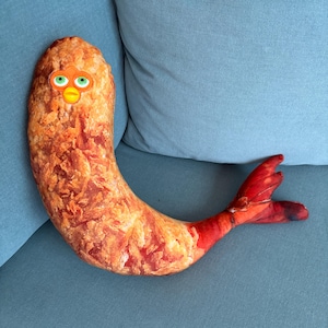 Può includere: Un peluche a forma di gambero fritto con una superficie impanata color marrone dorato, una coda rosso brillante e un volto in plastica con occhi verdi e un becco giallo inserito nella parte superiore del corpo ricurvo.