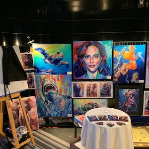 Puede incluir: Una colecci&oacute;n de pinturas coloridas expuestas en una exposici&oacute;n de arte. Las pinturas presentan una variedad de temas, incluyendo una tortuga, un tibur&oacute;n, un retrato de mujer, medusas y un pulpo. Las pinturas se exhiben en una pared blanca con un fondo negro.
