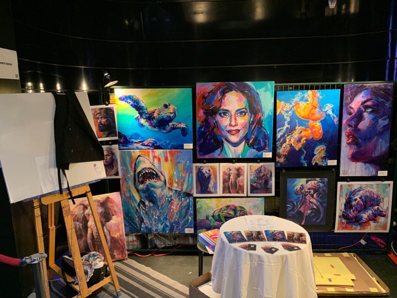 Puede incluir: Una colecci&oacute;n de pinturas coloridas expuestas en una exposici&oacute;n de arte. Las pinturas presentan una variedad de temas, incluyendo una tortuga, un tibur&oacute;n, un retrato de mujer, medusas y un pulpo. Las pinturas se exhiben en una pared blanca con un fondo negro.