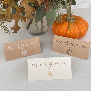 May include: Three place cards with the name "morgan" written in calligraphy. The place cards are decorated with fall-themed imagery, including a turkey, a pumpkin, and a leaf.