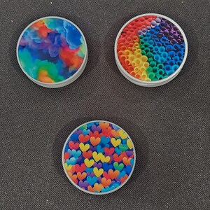 May include: Three round phone grips with colorful designs. One features a watercolor-like blend of blues, greens, oranges, and reds. Another has a rainbow of small tubes. The third has a pattern of colorful hearts.