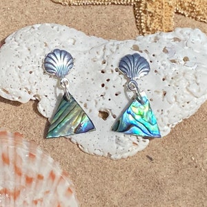 Puede incluir: Un par de pendientes de plata con un colgante de concha de abulón en forma de triángulo. Los pendientes tienen un pequeño encanto en forma de concha en la parte superior.