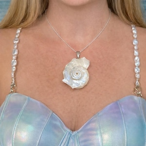 May include: A silver chain necklace featuring a large, iridescent white seashell pendant. The necklace is worn with a light blue, metallic-finish top that has straps decorated with white, irregular-shaped freshwater pearls on each side.