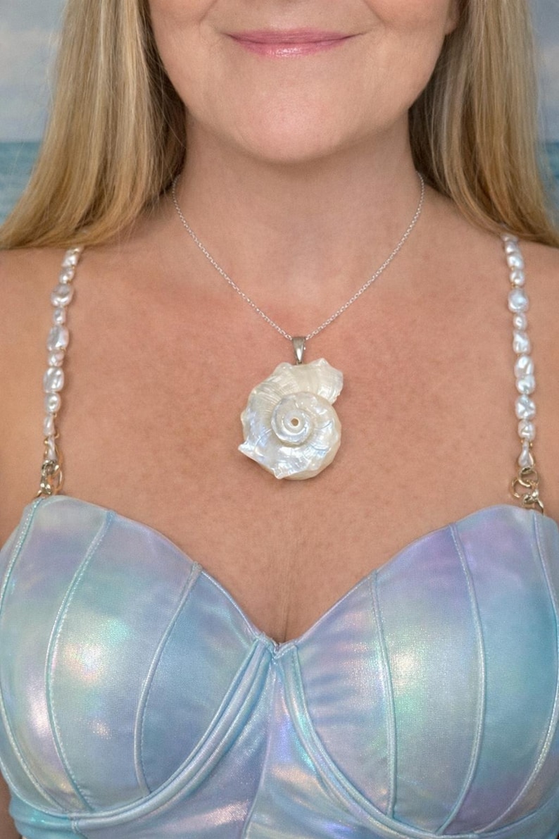 May include: A silver chain necklace featuring a large, iridescent white seashell pendant. The necklace is worn with a light blue, metallic-finish top that has straps decorated with white, irregular-shaped freshwater pearls on each side.