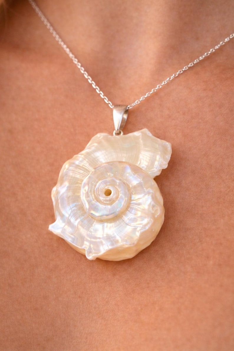May include: A pendant featuring a polished, iridescent white seashell with a spiral design, suspended from a thin silver chain. The shell displays a natural pearlescent sheen with subtle ridges and a smooth, curved texture throughout its surface.