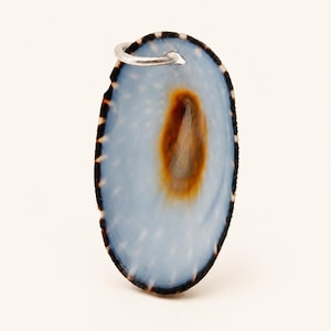Op de afbeelding: Een ovaalvormige hanger gemaakt van een natuurlijke schelp met een glad, lichtblauw oppervlak, een donkerbruine centrale vlek en een donkere, gespikkelde buitenrand. Een kleine, zilverkleurige metalen ring is aan de bovenkant bevestigd.