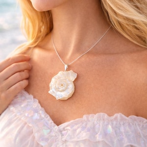 May include: A silver chain necklace featuring a large, iridescent white seashell pendant with a spiral center. The shell has a pearlescent finish and is attached to the chain with a simple silver bail, resting against the collarbone area.
