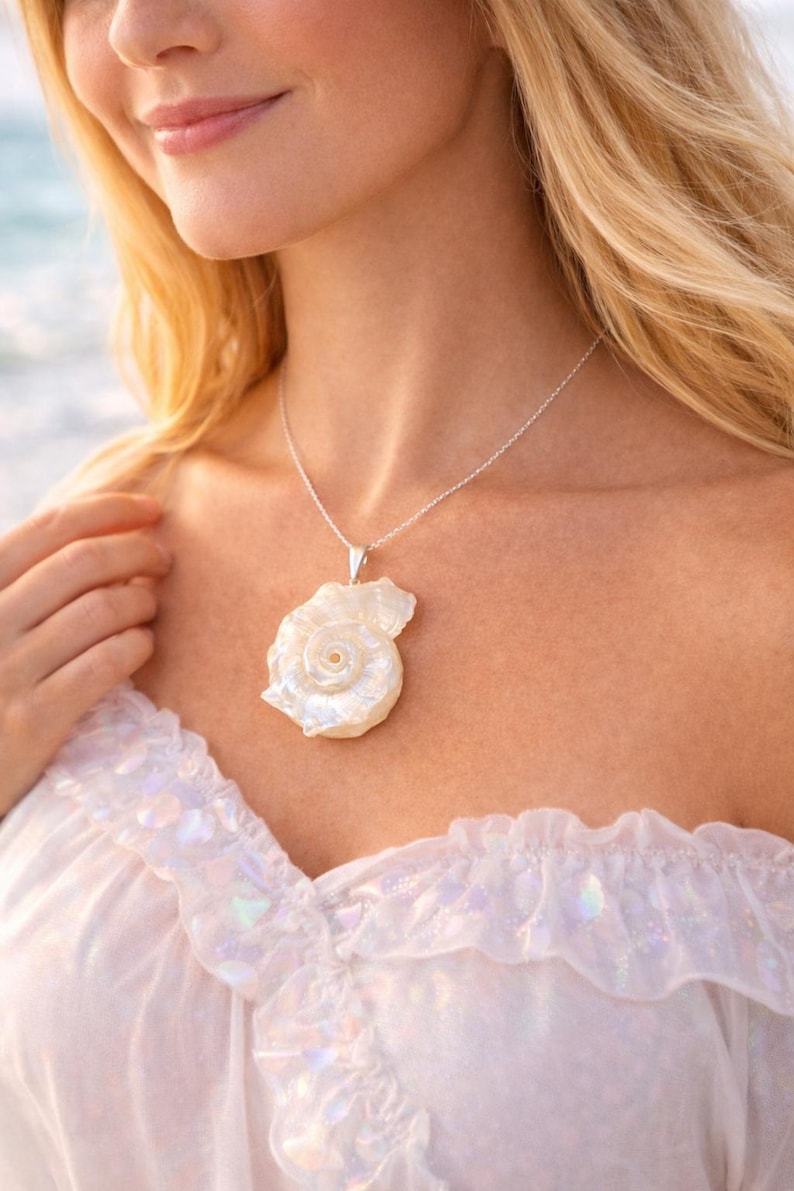 May include: A silver chain necklace featuring a large, iridescent white seashell pendant with a spiral center. The shell has a pearlescent finish and is attached to the chain with a simple silver bail, resting against the collarbone area.