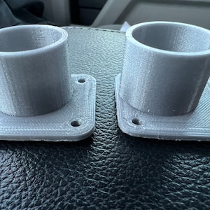 May include: Two gray 3D printed plastic cup holders with square bases. Each base has four holes for mounting.